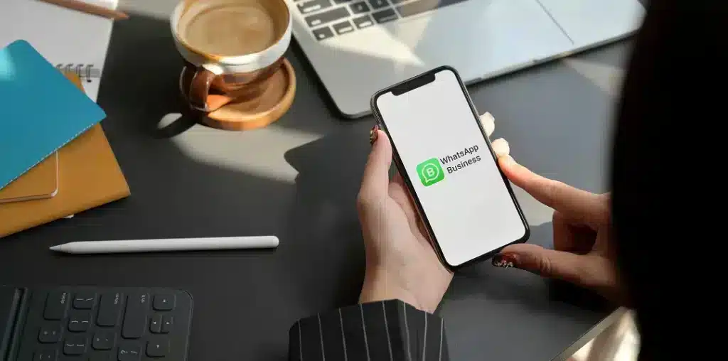 Persona sosteniendo un teléfono con WhatsApp Business en pantalla, sobre escritorio con laptop y café