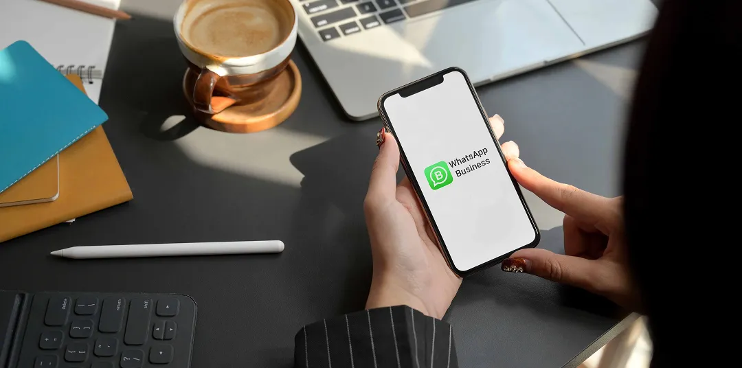 Persona sosteniendo un teléfono con WhatsApp Business en pantalla, sobre escritorio con laptop y café